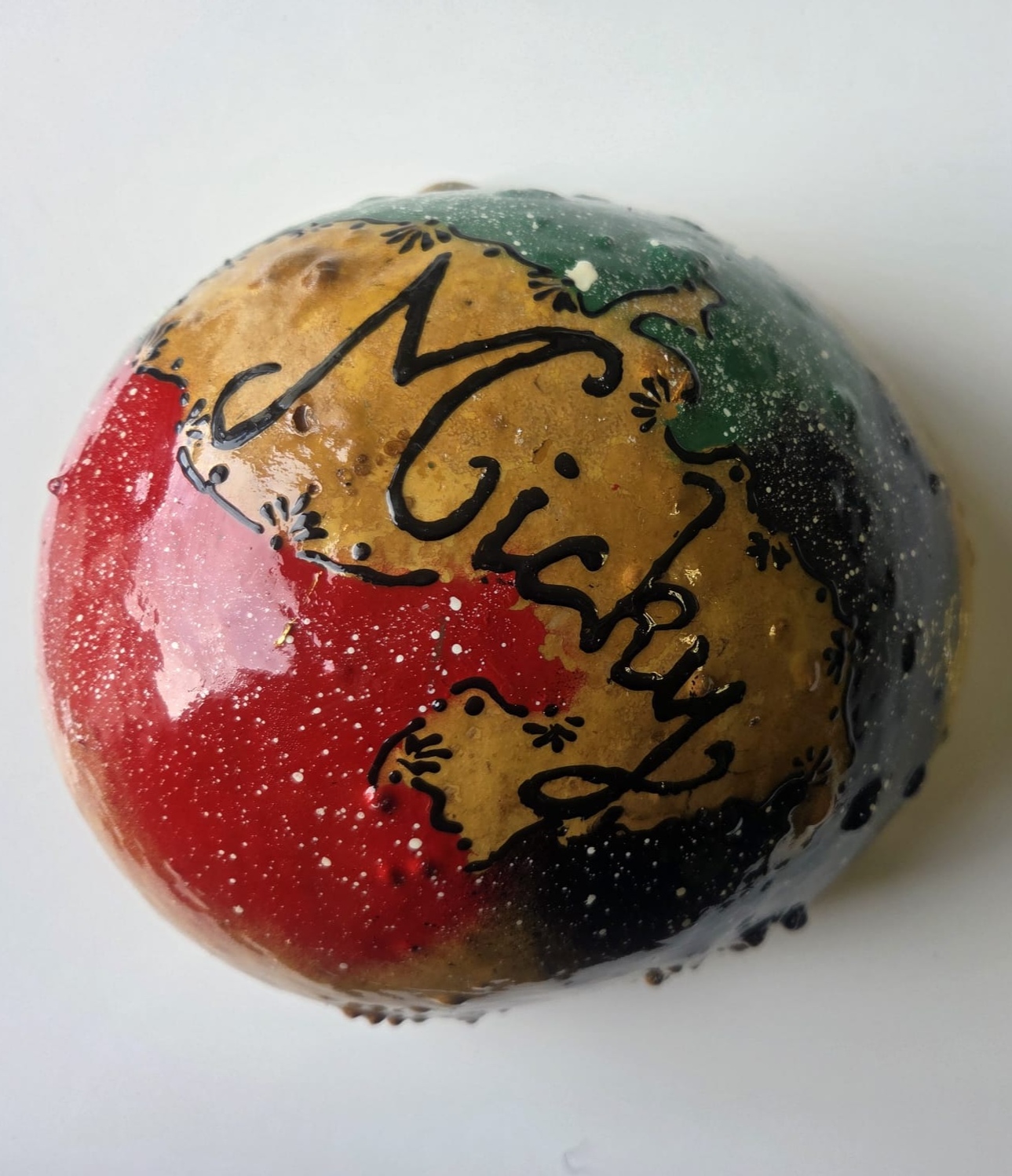Un dessert brillant en forme de dôme présente des sections rouges, dorées, vertes et noires avec le nom Micky écrit en cursive sur le dessus, le tout sur un fond blanc uni.